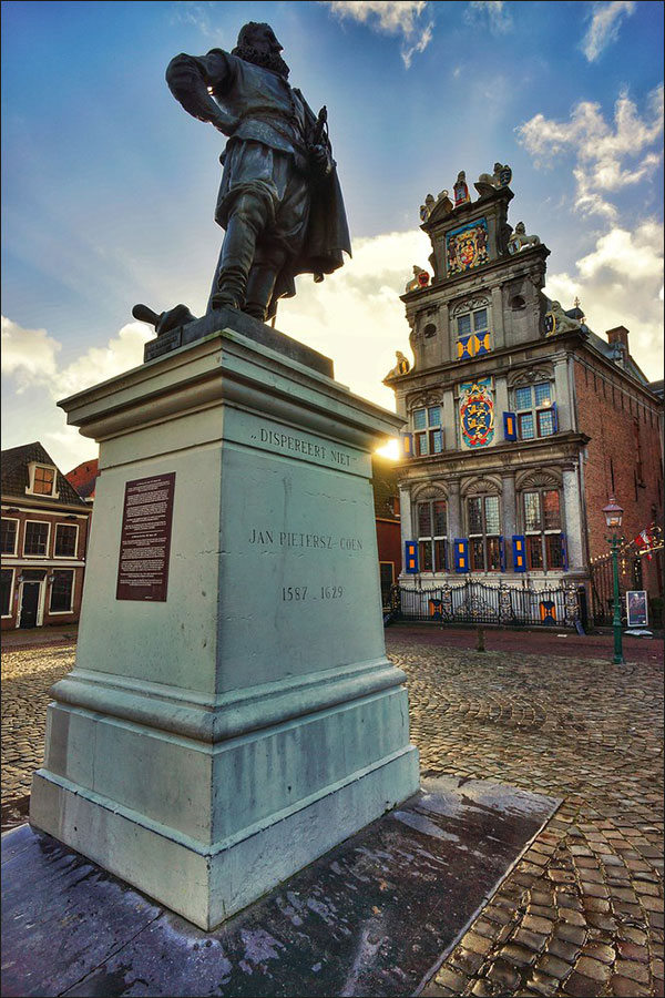Het Rijksmonumentale standbeeld van Jan Pieterszoon Coen in Hoorn, tegen een diepblauwe lucht met witte wolken. Op de achtergrond straalt het Statencollege, een gebouw uit 1632 met een pronkgevel in Hollandse Renaissancestijl. Het wapen van West-Friesland, met de hoogte van een volle verdieping, staat in de gevel centraal.